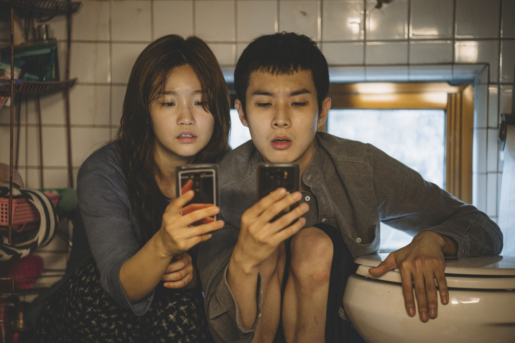 Two young people look at their phones while crouching down and leaning against a toilet